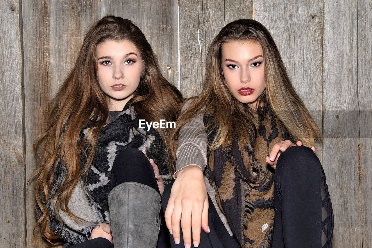 Portrait of sisters wearing warm clothing while sitting against wooden wall