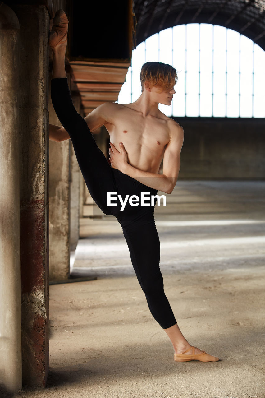 Amazing young gymnast in black leotard and pointe shoes doing longitudinal twine putting foot on wall near window in room and looking at camera