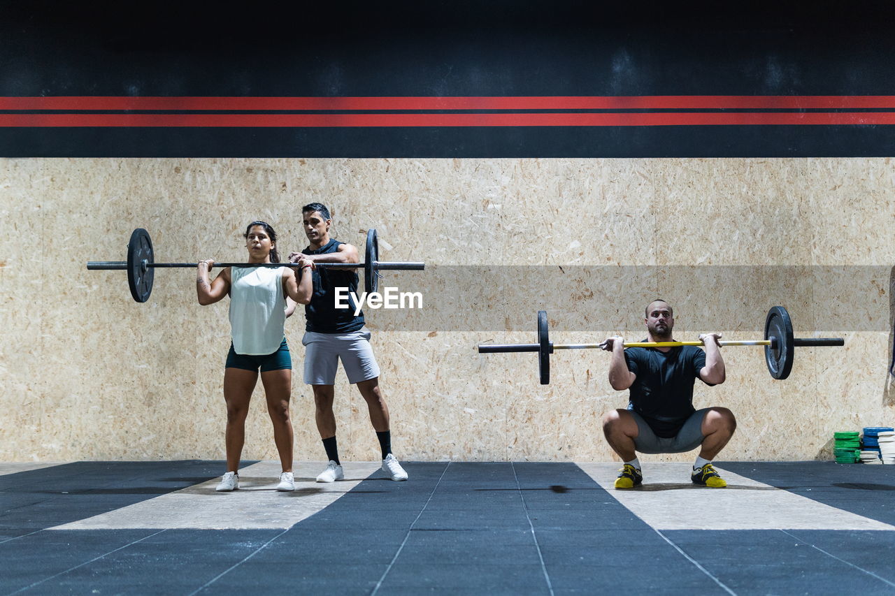 Full body group of strong hispanic athletes lifting heavy barbells near wall in light gym during weightlifting training together