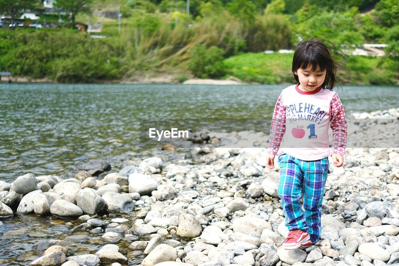one person, childhood, child, water, full length, casual clothing, nature, rock, standing, female, stone, innocence, cute, day, women, toddler, front view, leisure activity, smiling, lake, emotion, lifestyles, outdoors, clothing, pebble, person, portrait, happiness, plant, tree, looking at camera, black hair, land, looking, beauty in nature