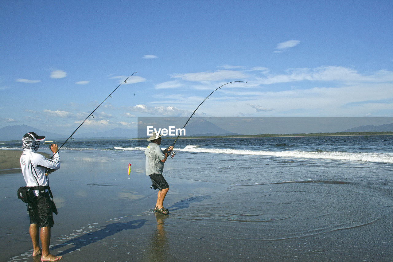 REAR VIEW OF MEN FISHING IN FRONT OF SEA AGAINST SKY