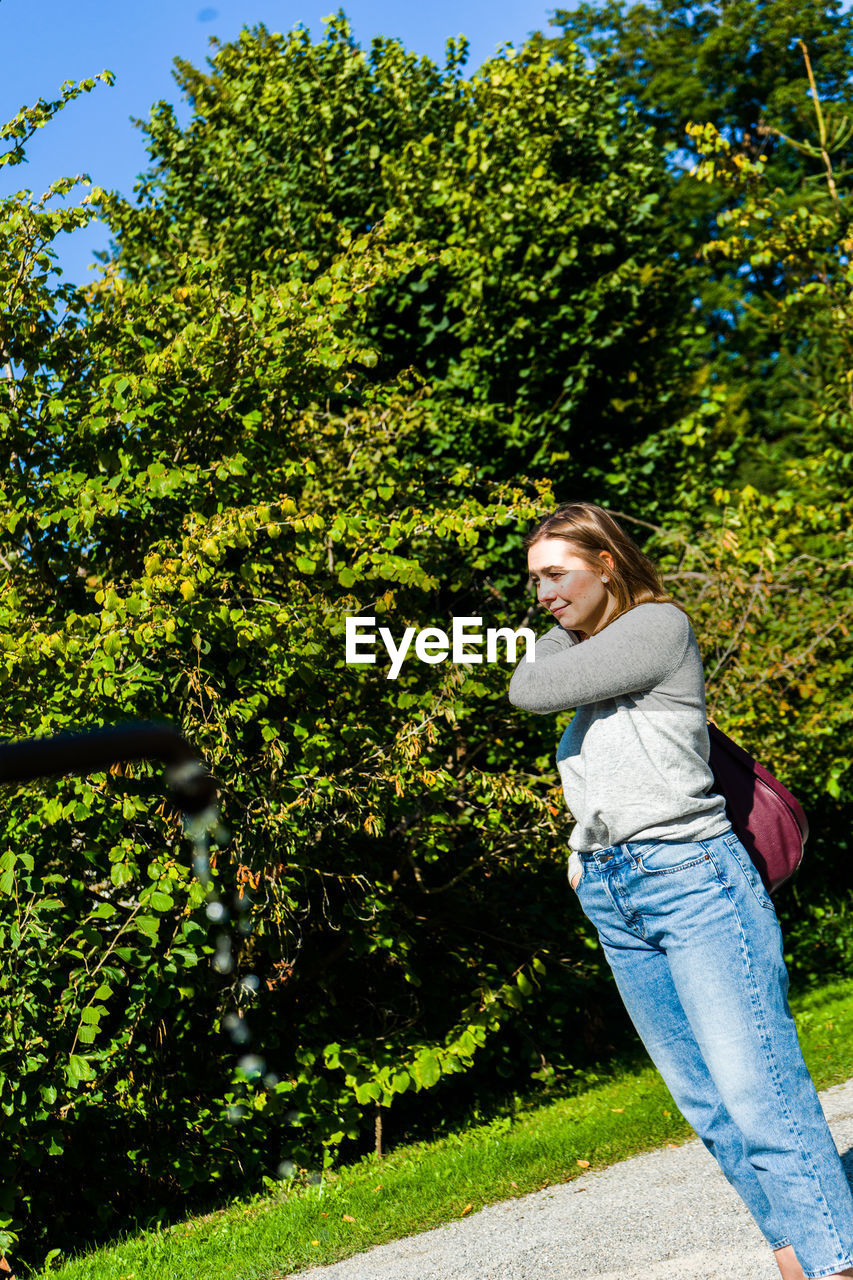 SMILING WOMAN STANDING BY PLANTS AGAINST TREES