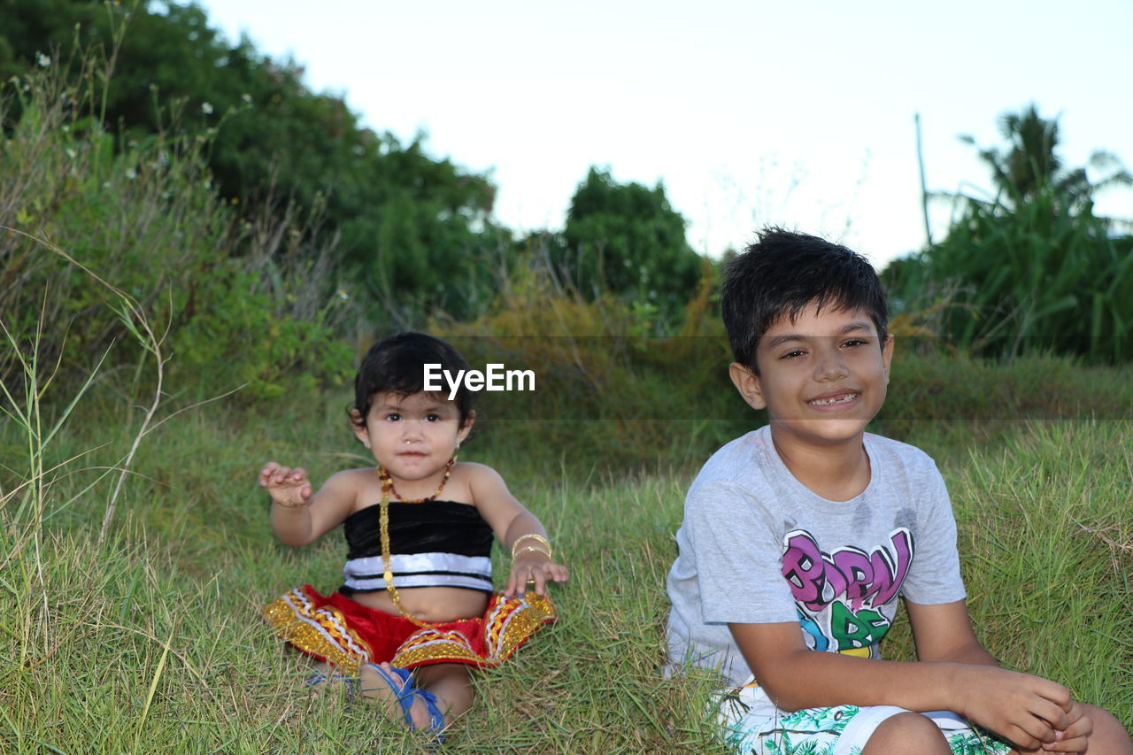 Portrait of cute siblings sitting on grass against sky