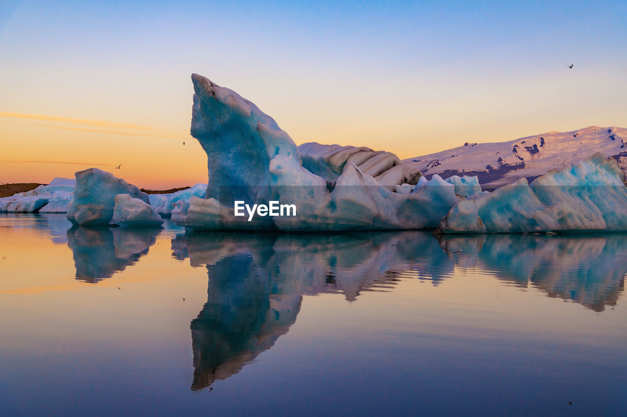 Scenic view of icebergs in sea against snowcapped mountains and sky during sunset