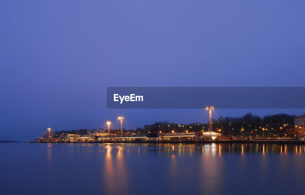 Illuminated city by sea against clear blue sky at night