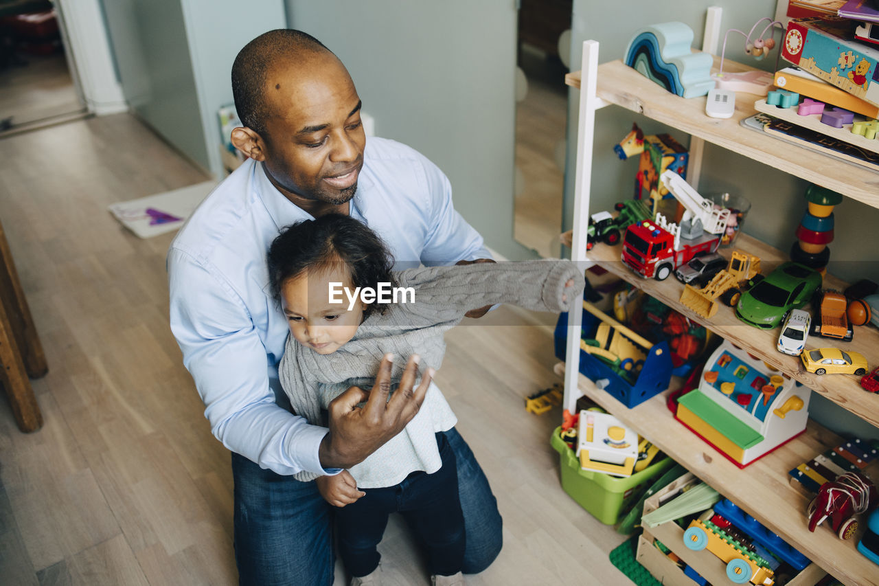 High angle view of father dressing daughter while kneeling by toys on shelf at home