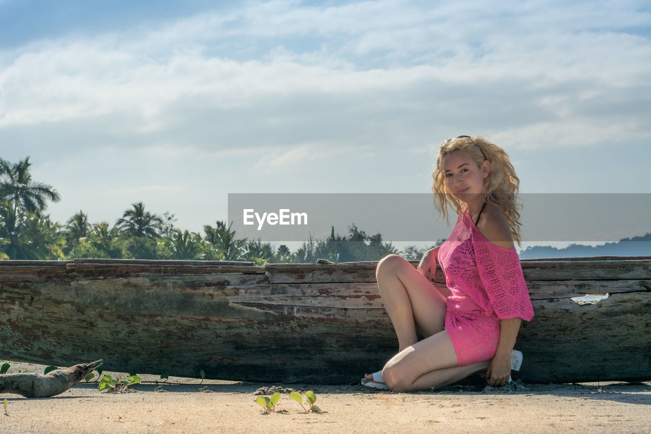 Portrait of beautiful woman kneeling by weathered wooden boat on shore at beach against sky