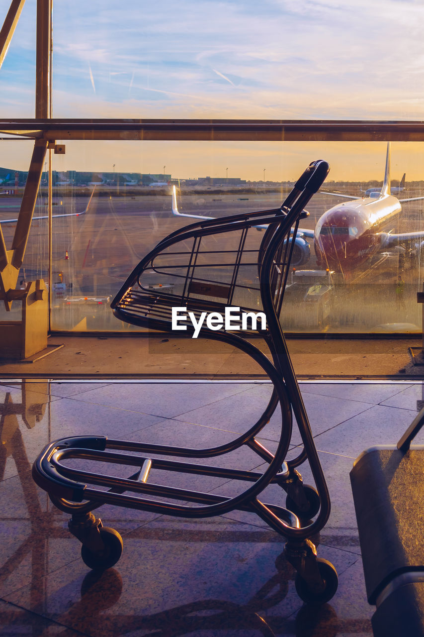 Close-up of cart by window with airplane on runway against sky