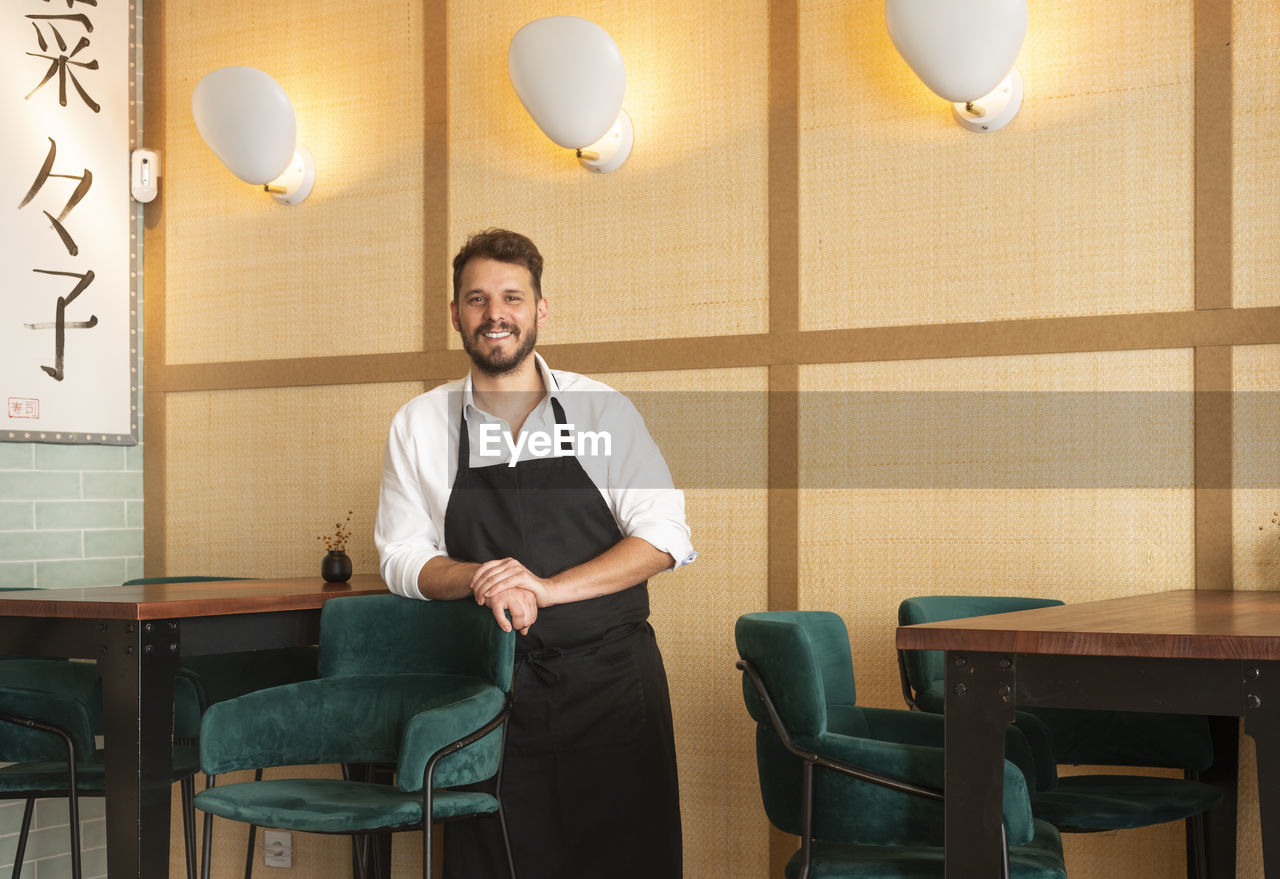Cheerful male chef in apron standing in sushi restaurant and looking at camera