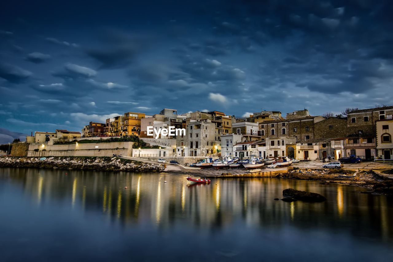 Buildings by river against cloudy sky in city during night