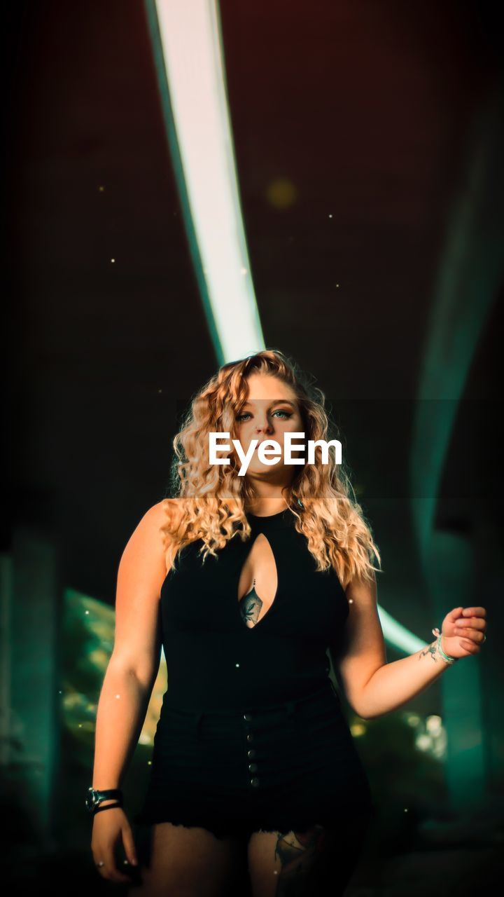 PORTRAIT OF A BEAUTIFUL YOUNG WOMAN STANDING IN ILLUMINATED LIGHT