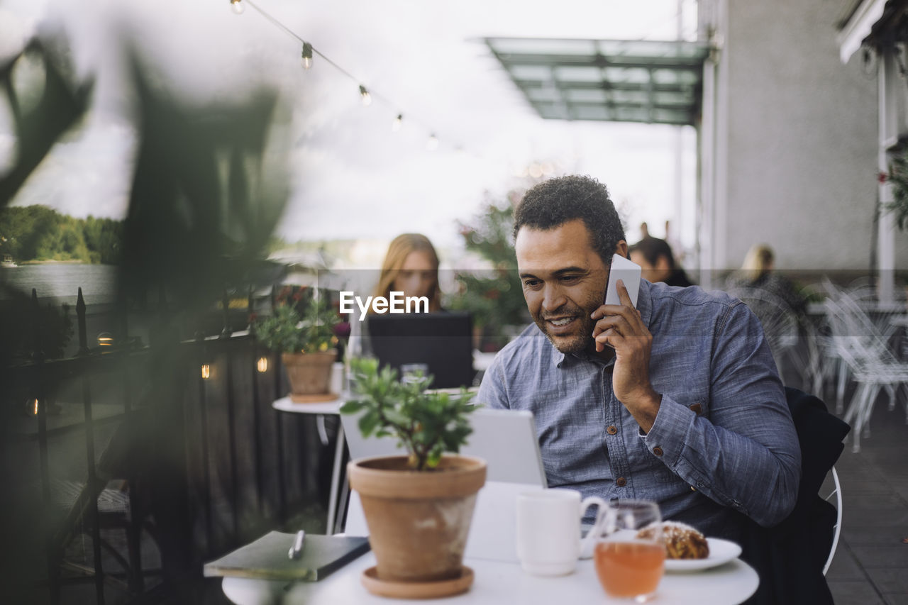 Smiling male entrepreneur talking on smart phone while sitting with laptop at cafe