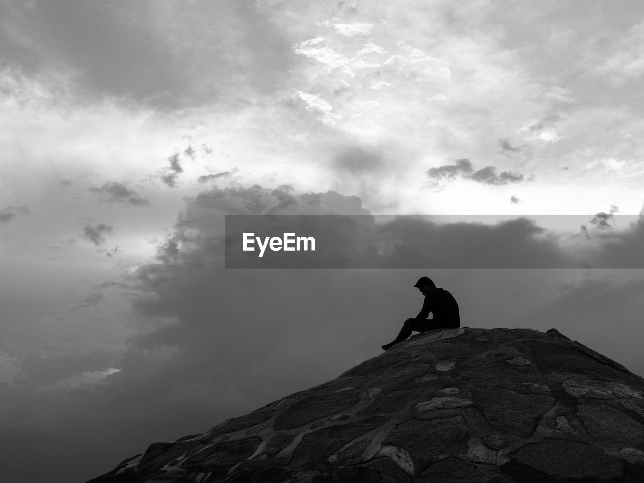 Silhouette of man sitting in thinking pose on breakwater under dramatic cloudy sky.