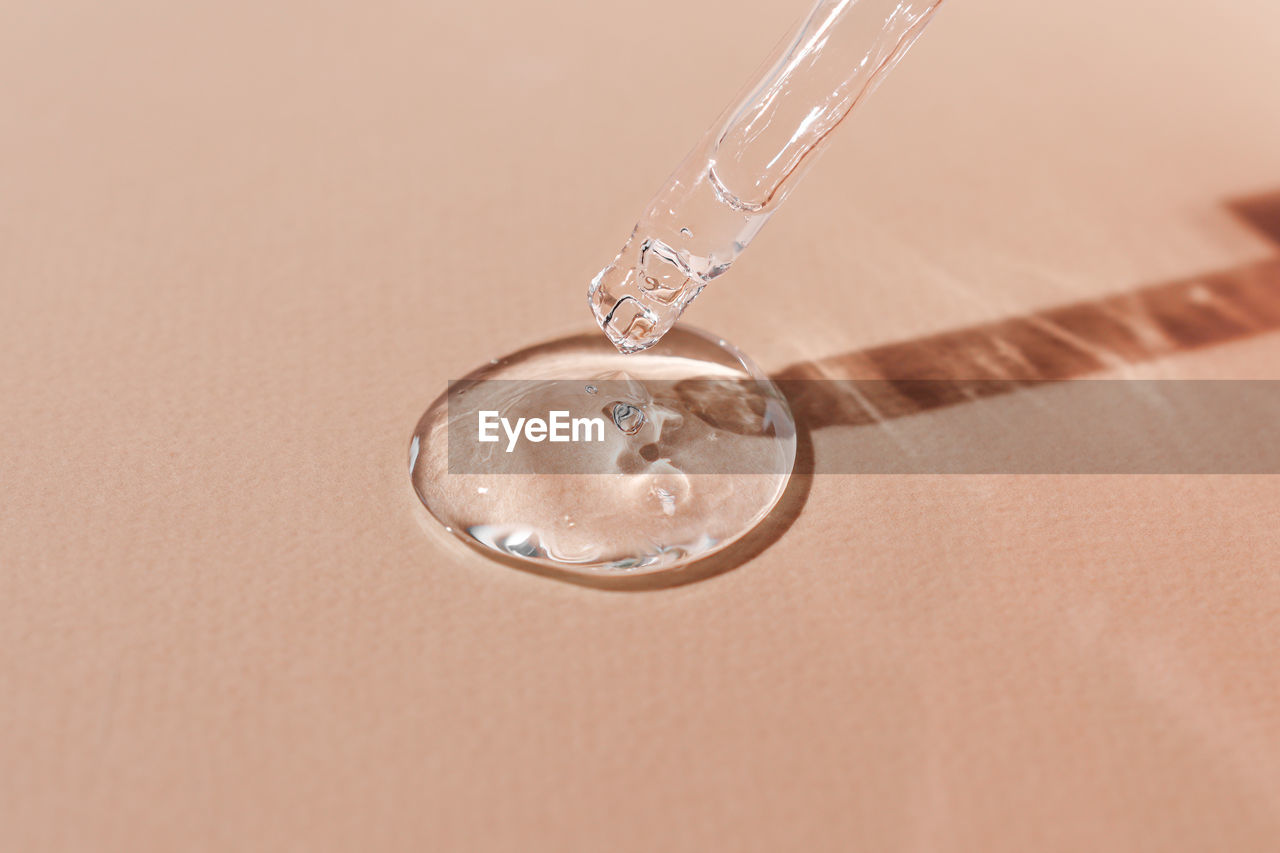 A sample of transparent hyaluronic acid on a beige background flows down from a glass pipette.