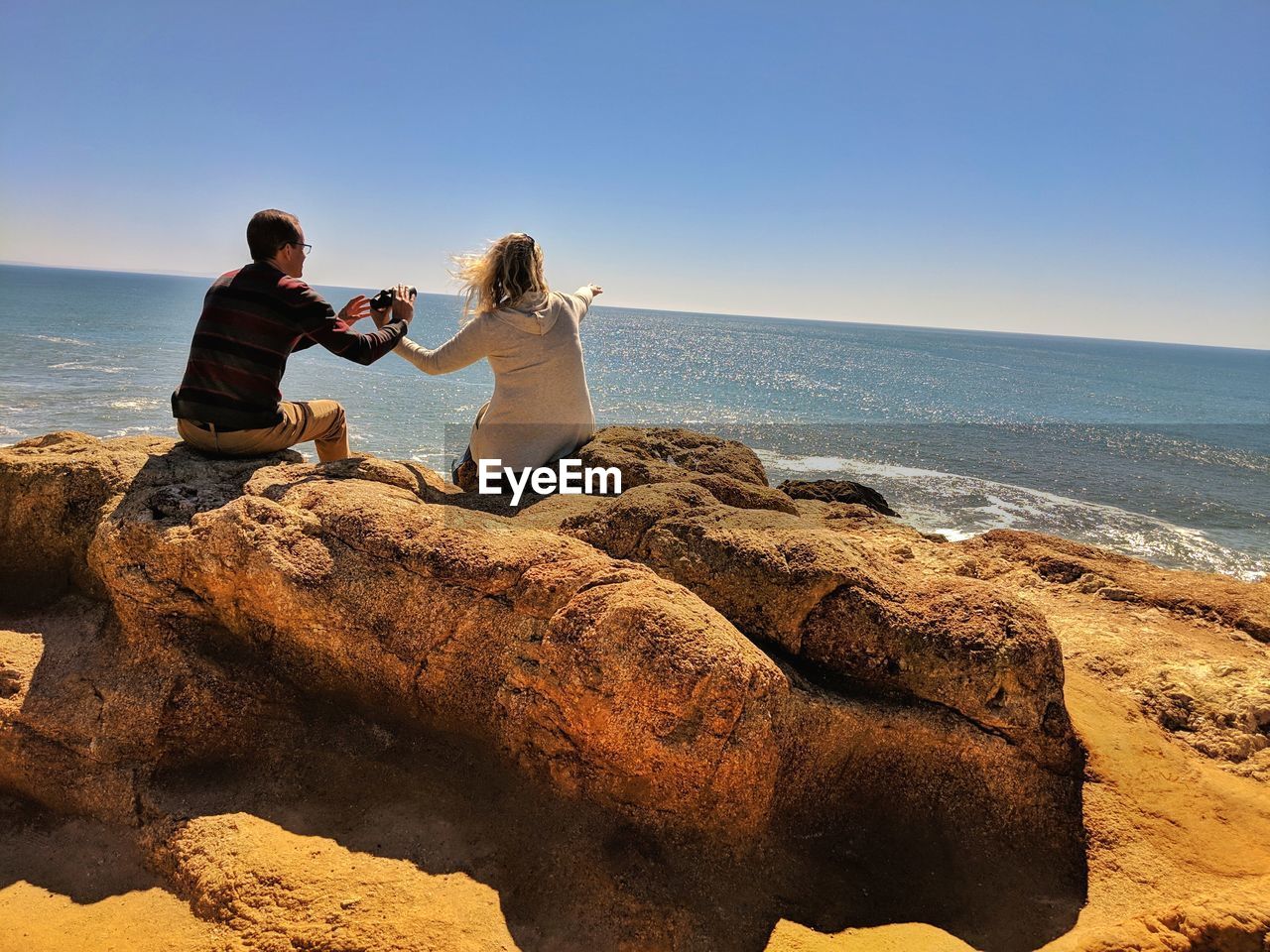 Couple on sandstone boulders at ocean whale watching woman pointing out to sea.