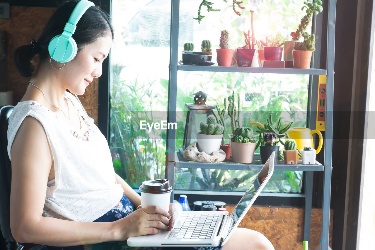 Side view of woman using laptop while sitting against potted plants on shelves at home