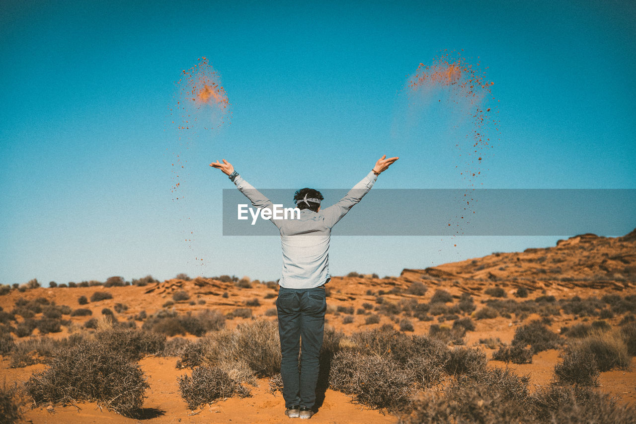 Rear view of man throwing sand while standing at desert against sky