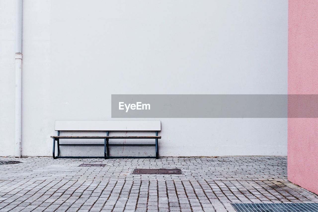 Empty bench on sidewalk against white wall