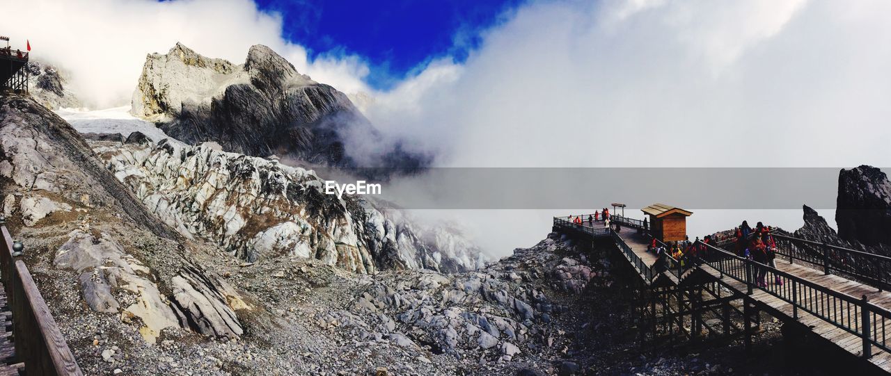 People on walkway by rocky mountains against cloudy sky during winter