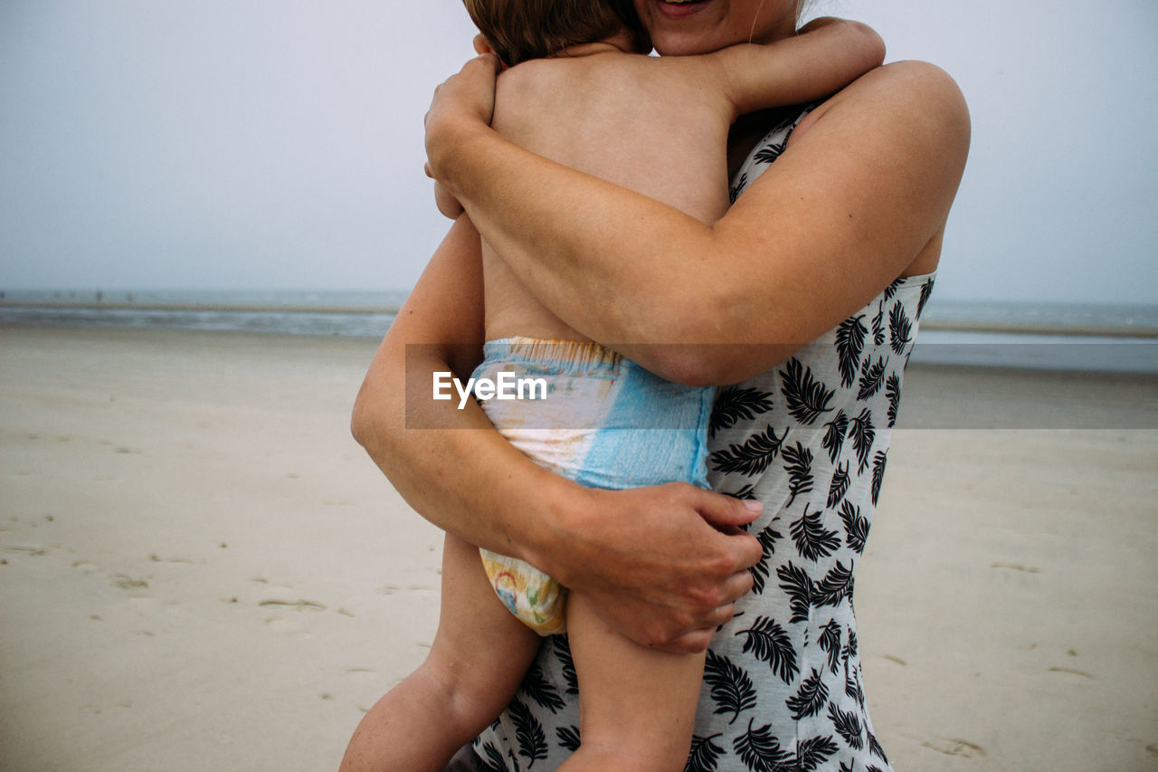 Midsection of woman holding her baby and standing at beach against sky.