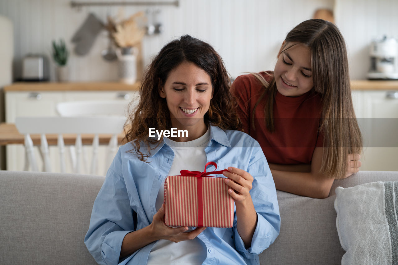 Excited young woman mother getting wrapped gift box present from daughter while sitting on sofa