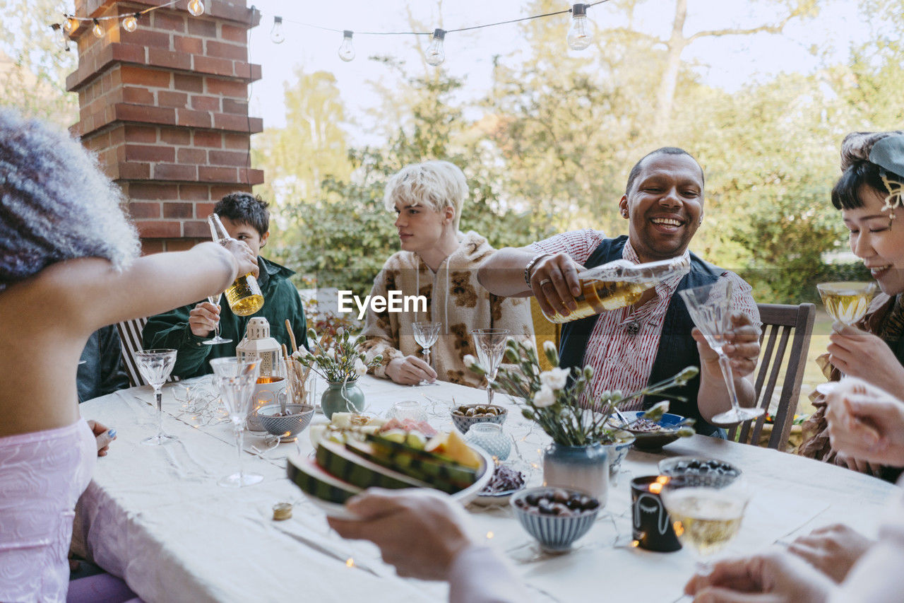 Happy friends of lgbtq community drinking wine while sitting at table during dinner party in back yard