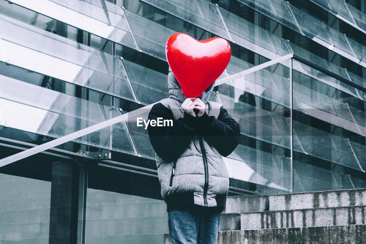 Boy covering face with red heart shape helium balloon against building in city