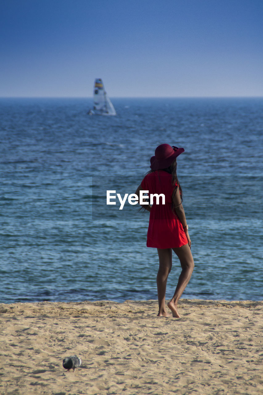 Rear view of woman in red dress standing at beach against blue sky
