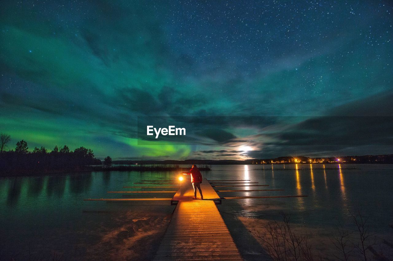 Rear view of person standing on pier over lake at night