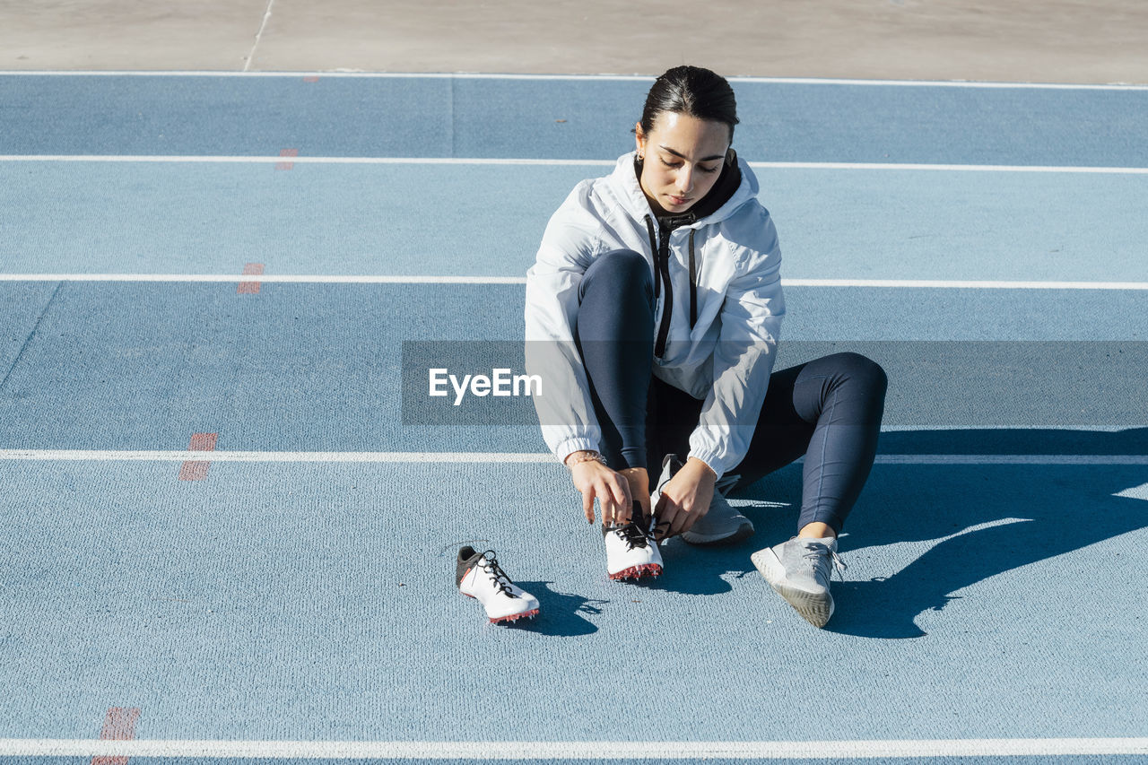 Female athlete in sportswear sitting on racetrack and changing footwear during track and field workout on sunny day on stadium