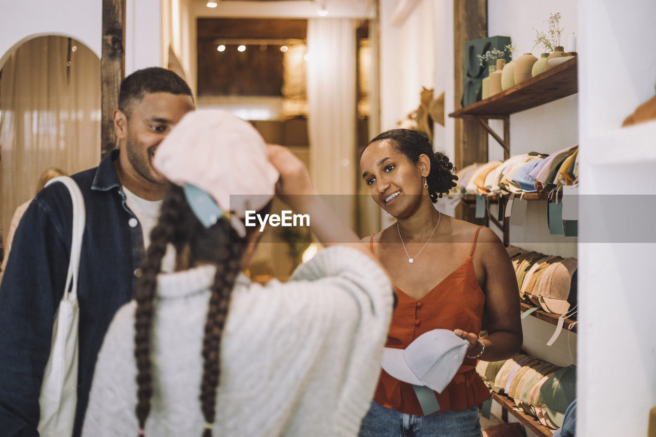 Smiling saleswoman and man looking at girl wearing cap in fashion clothing store