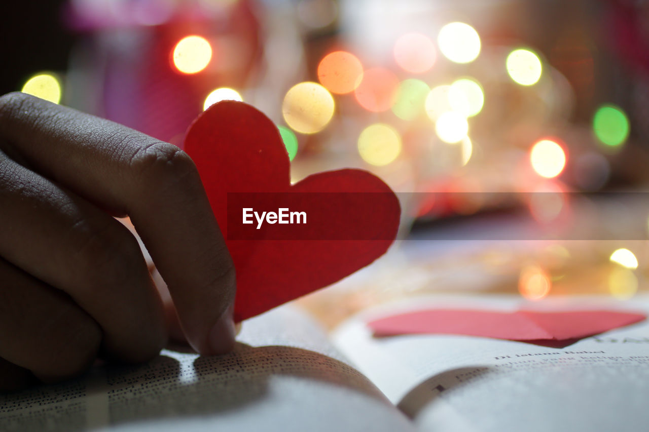 Close-up of hand holding red heart shape on book. 