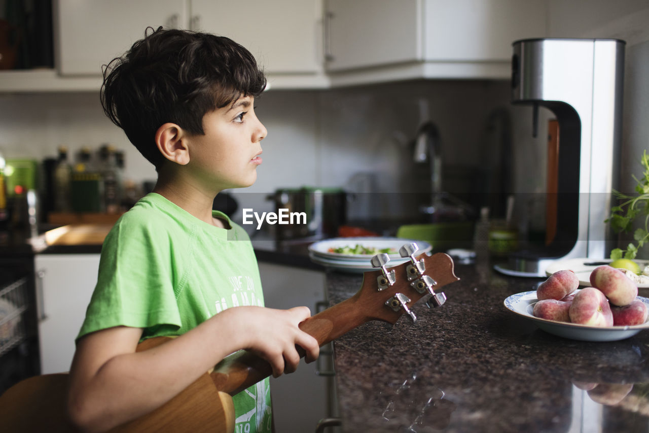 Thoughtful boy holding guitar while standing at kitchen counter