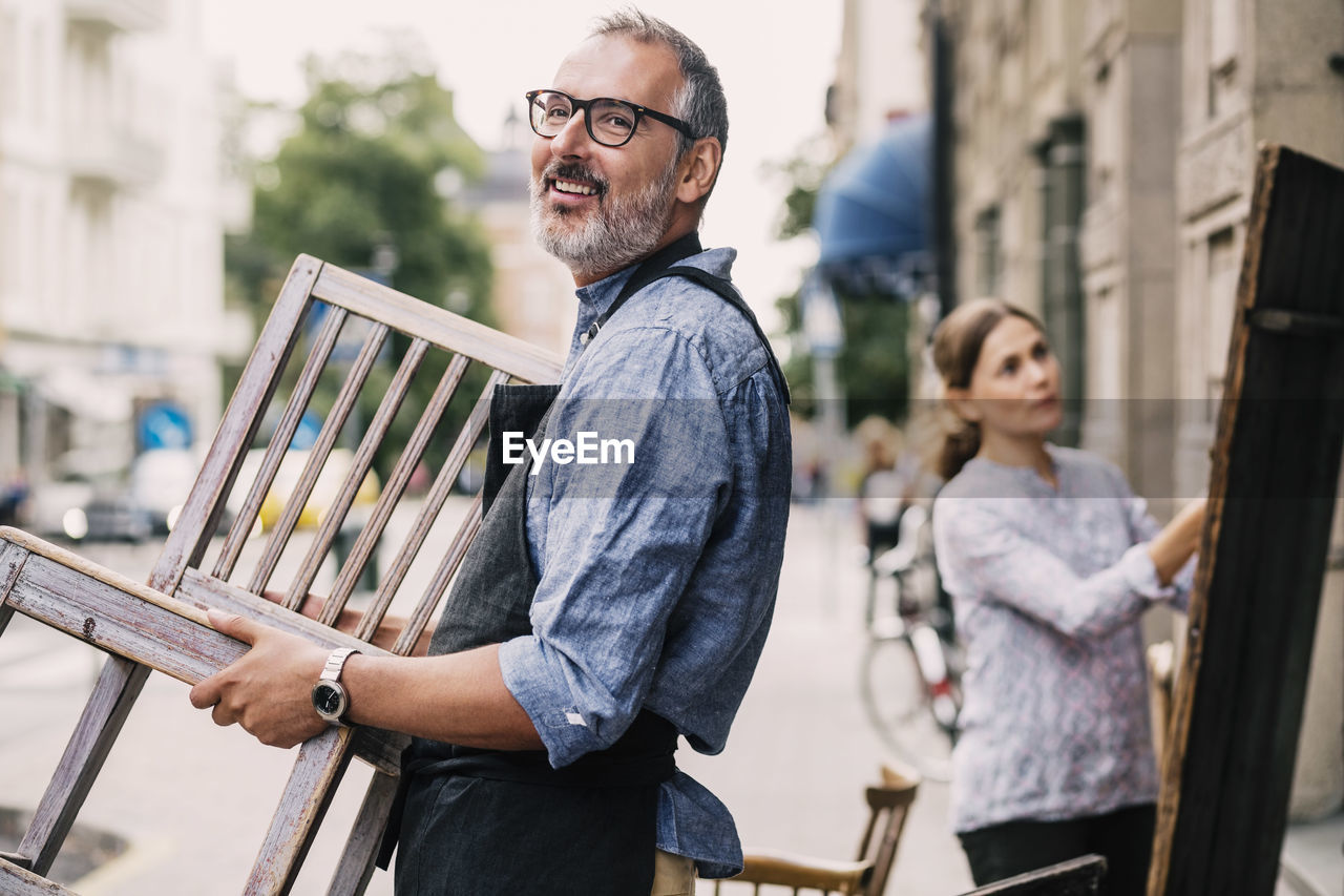 Male owner carrying chair while colleague working in background outside antique shop