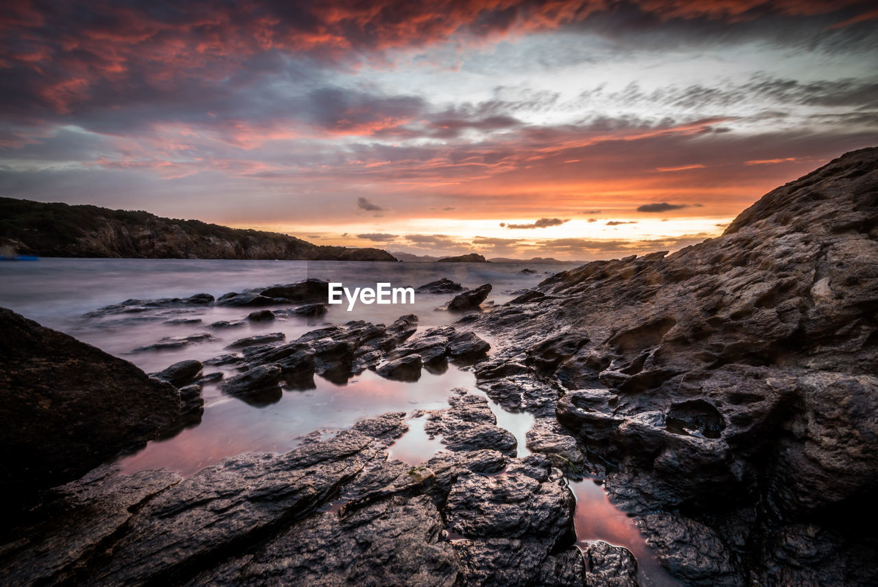 SCENIC VIEW OF ROCKS AT SEA AGAINST SKY DURING SUNSET