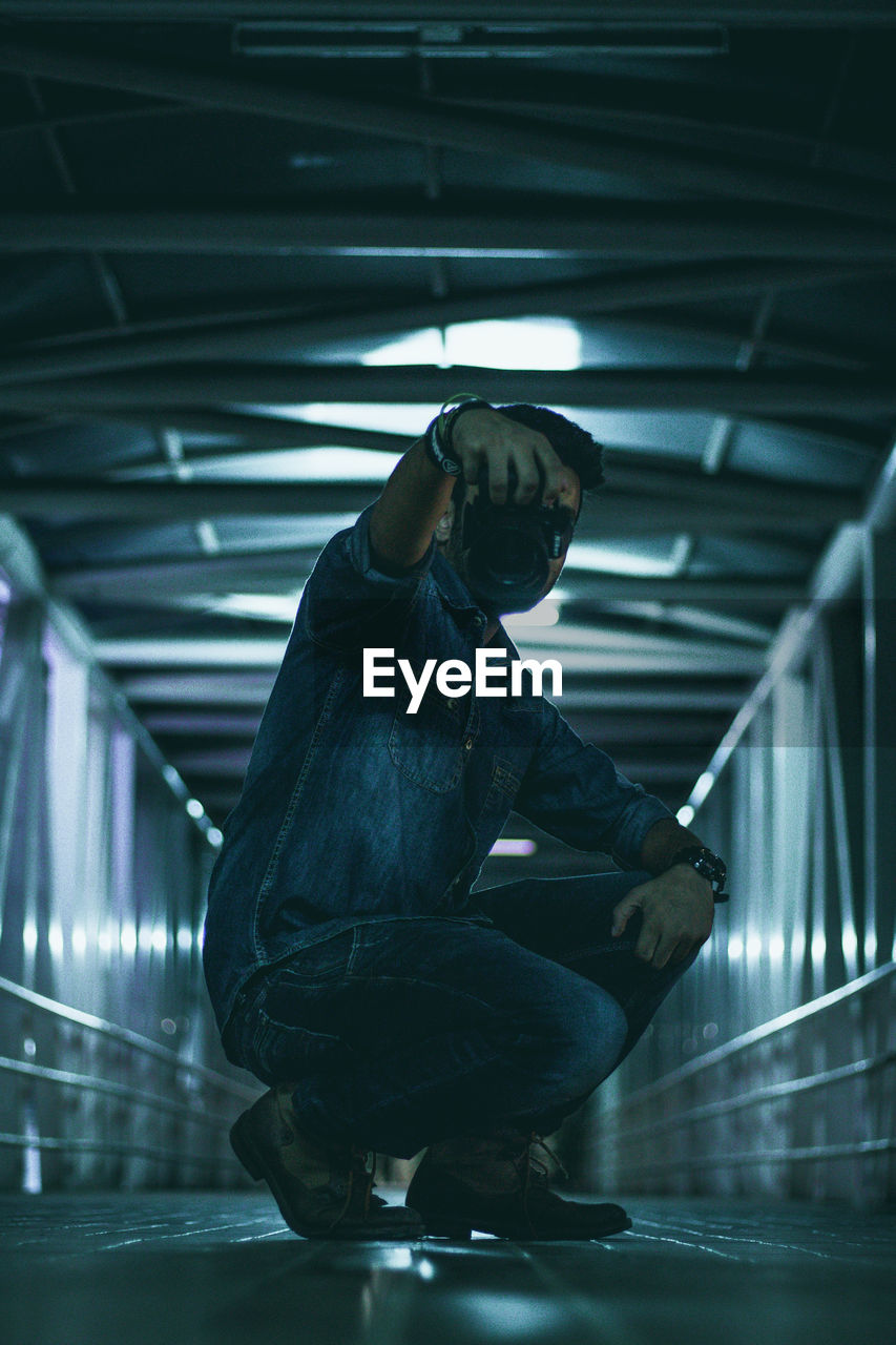 Full length side view of man holding camera while crouching on bridge