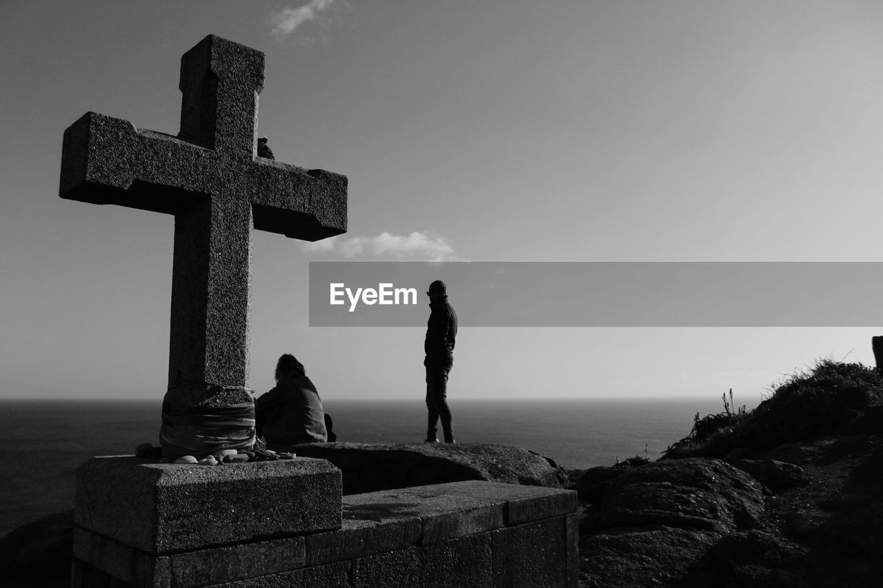 People on rock by religious cross against sky