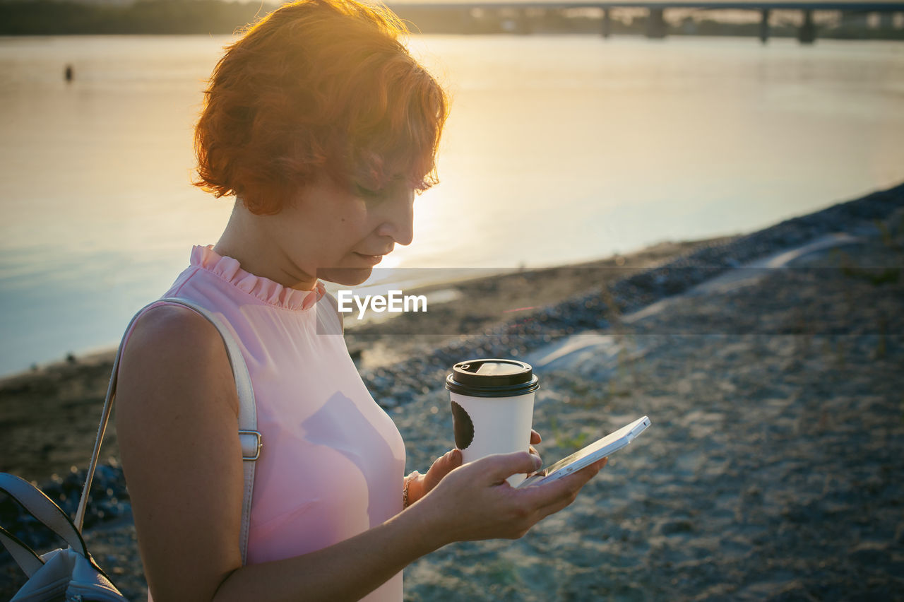 Mid adult woman using mobile phone while standing by river against sky during sunset