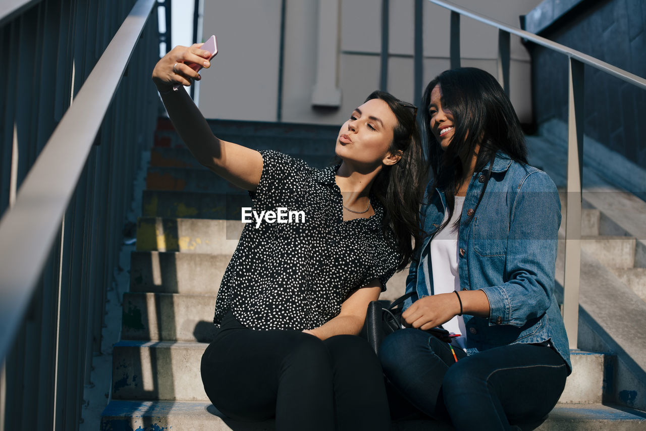 Low angle view of young woman taking selfie with female friend while sitting on staircase