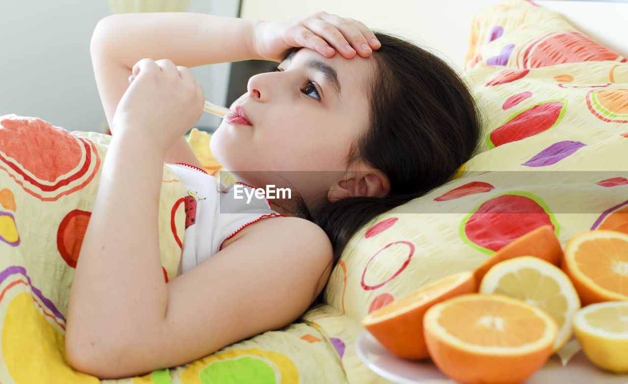 Teenage girl holding thermometer in mouth while lying on bed at home