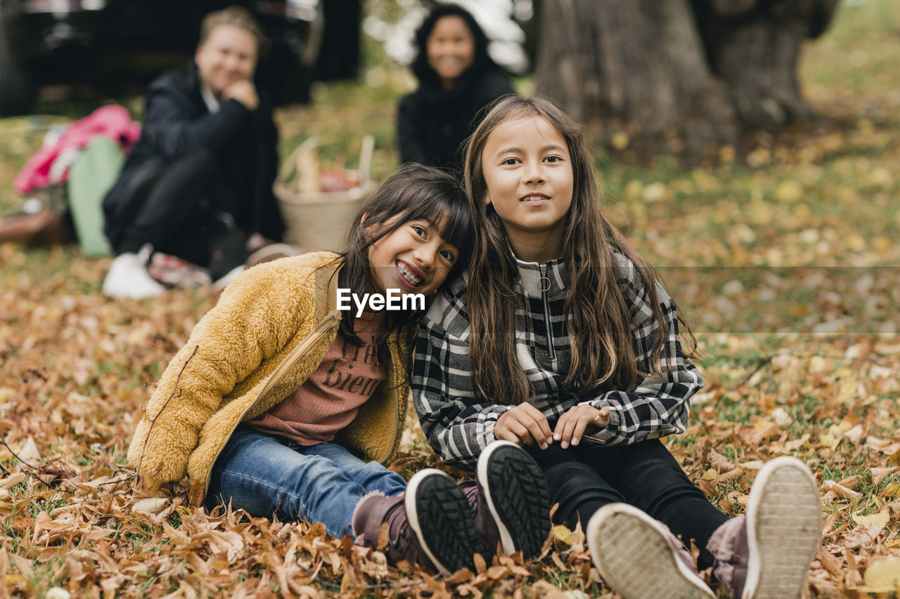 Portrait of smiling sisters sitting on autumn leaves during picnic with parents