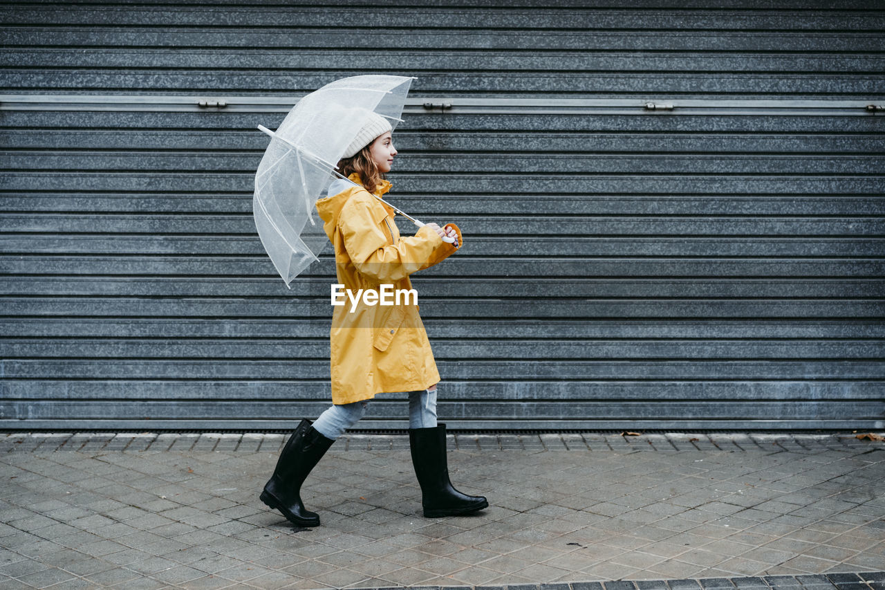Girl wearing raincoat and jump boot holding umbrella while walking on sidewalk