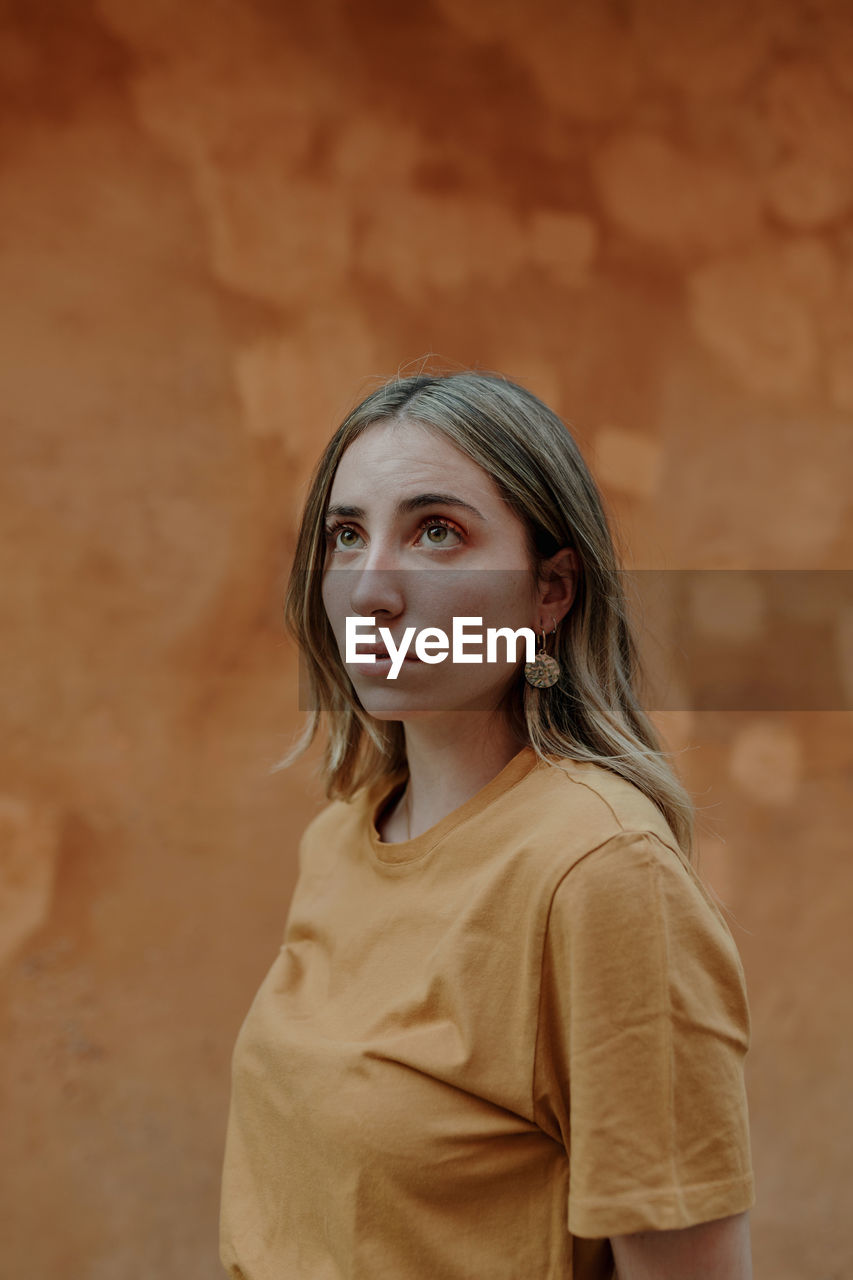 Young content blonde female in casual wear with wavy hair looking away while standing on brown background in town