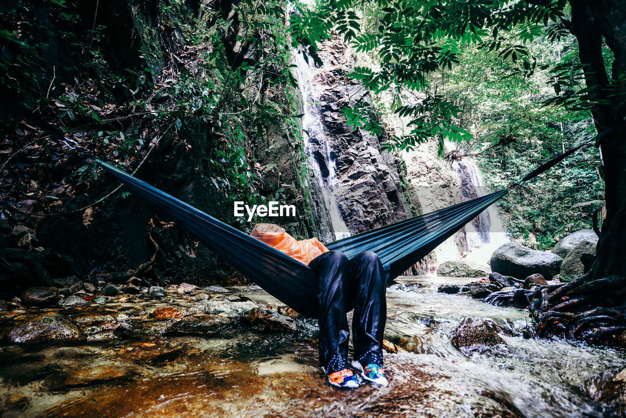 Young woman resting on hammock against waterfall in forest