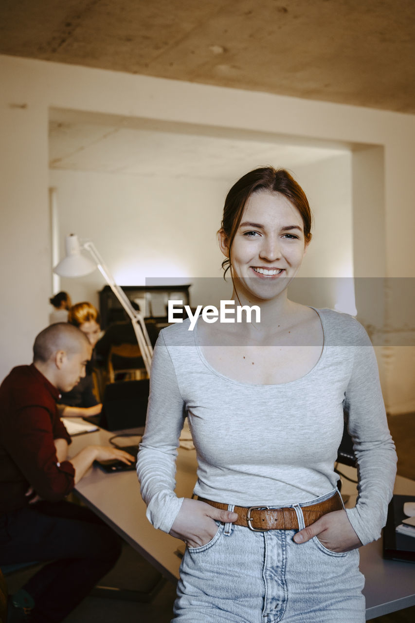 Portrait of smiling businesswoman with hands in pockets while colleague working at desk in office