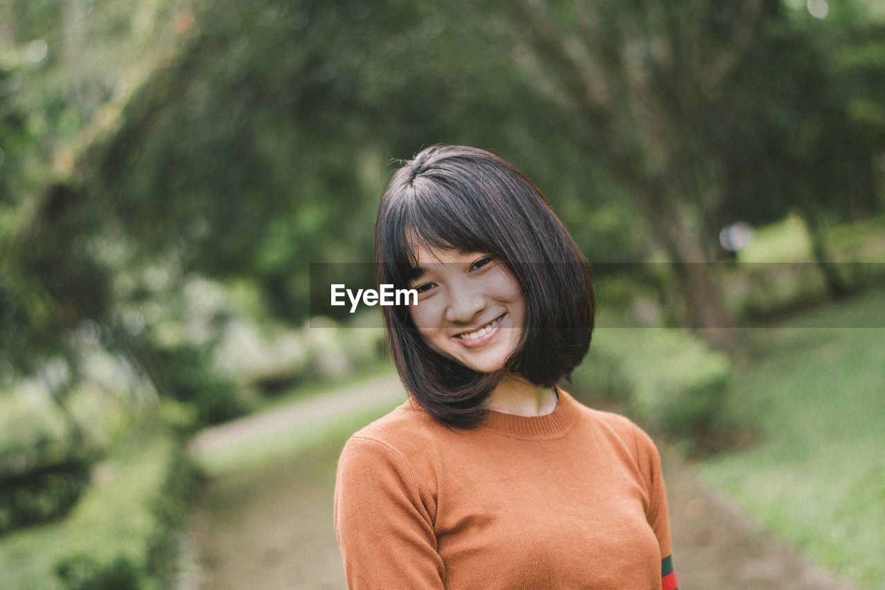 Portrait of smiling young woman standing against trees in park