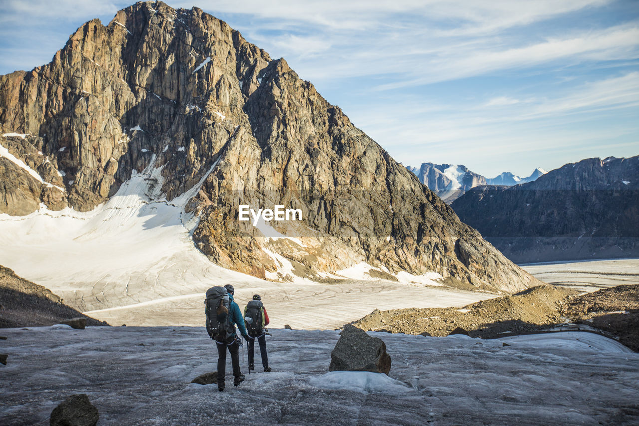 Two backpackers cross a glacier below mountain range.