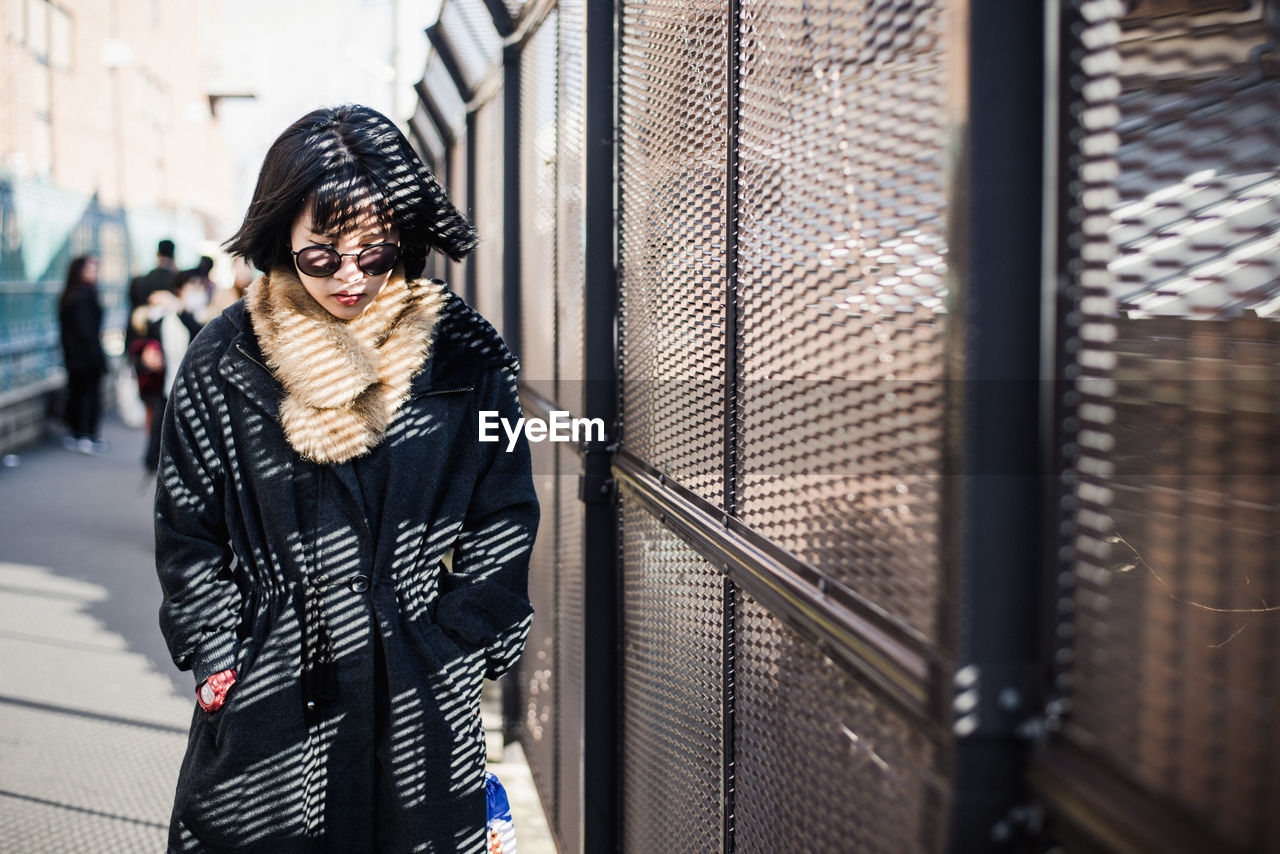 Young woman in warm clothing wearing sunglasses standing by built structure