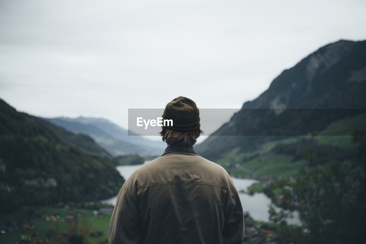 Rear view of man standing near lake and mountains against clear sky