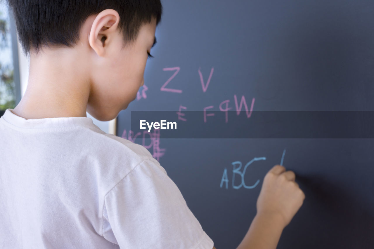 Rear view of boy writing with chalk on blackboard
